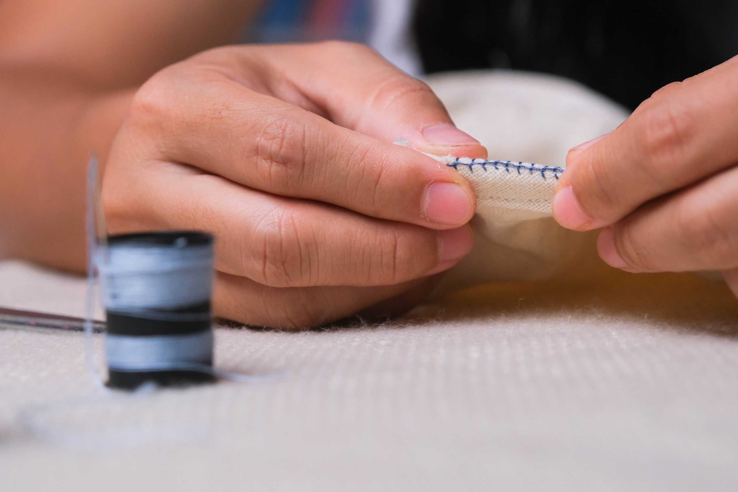 Hand of a seamstress at work with cloth fabric. Mother sews a beautiful dress for her little daughter by hand, close-up. Woman hand sews seams with blue threads.